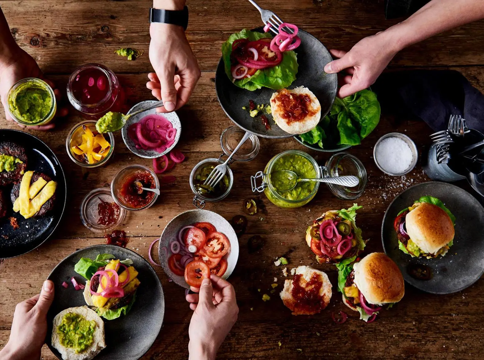 Vários pratos de comida vegana em cima da mesa, algumas mãos estão pegando alimentos deles.