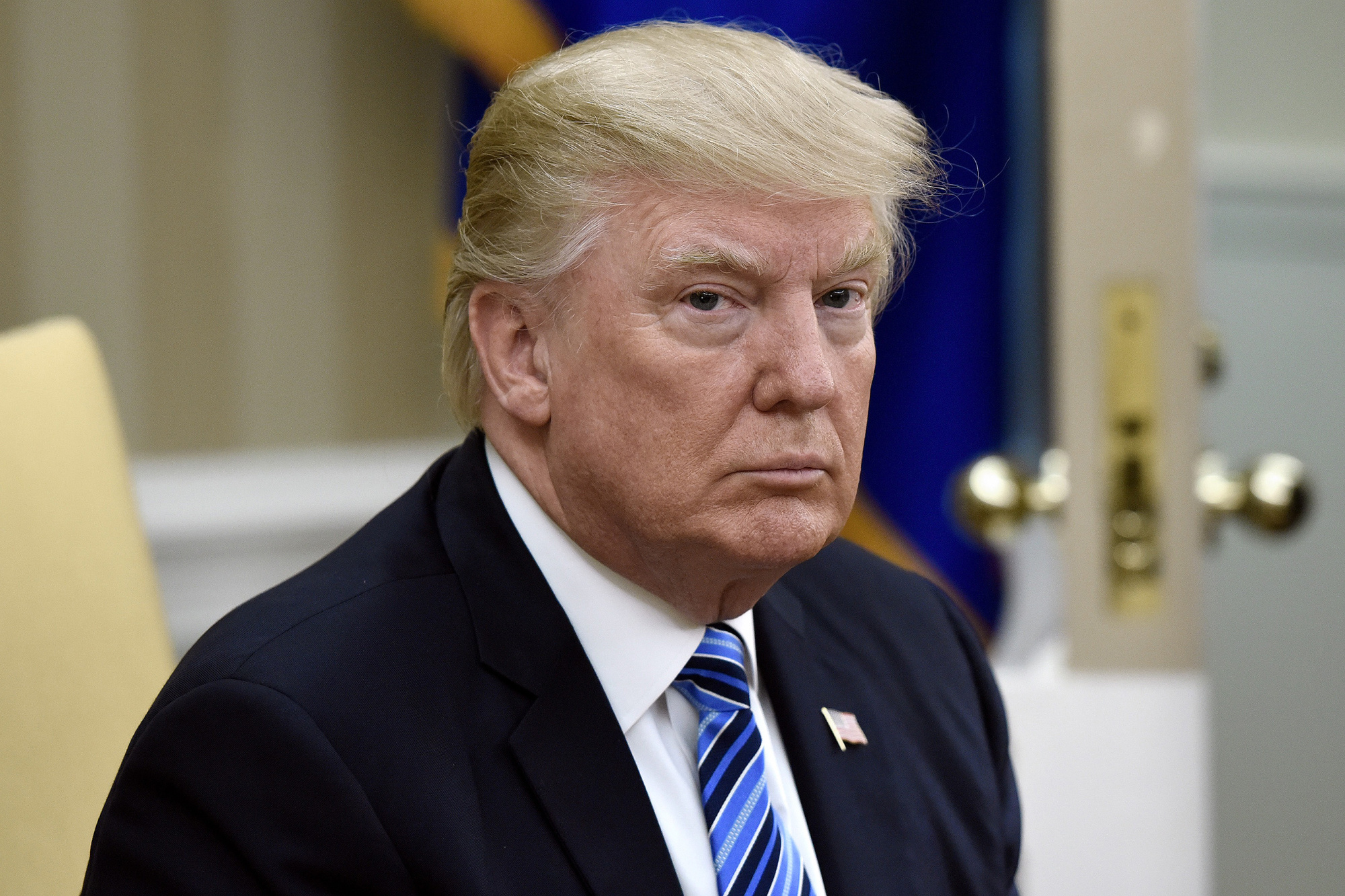 Donald Trump usando um terno preto, camisa branca e gravata azul com listras. Na sua lapela direita, um broche da bandeira dos Estados Unidos